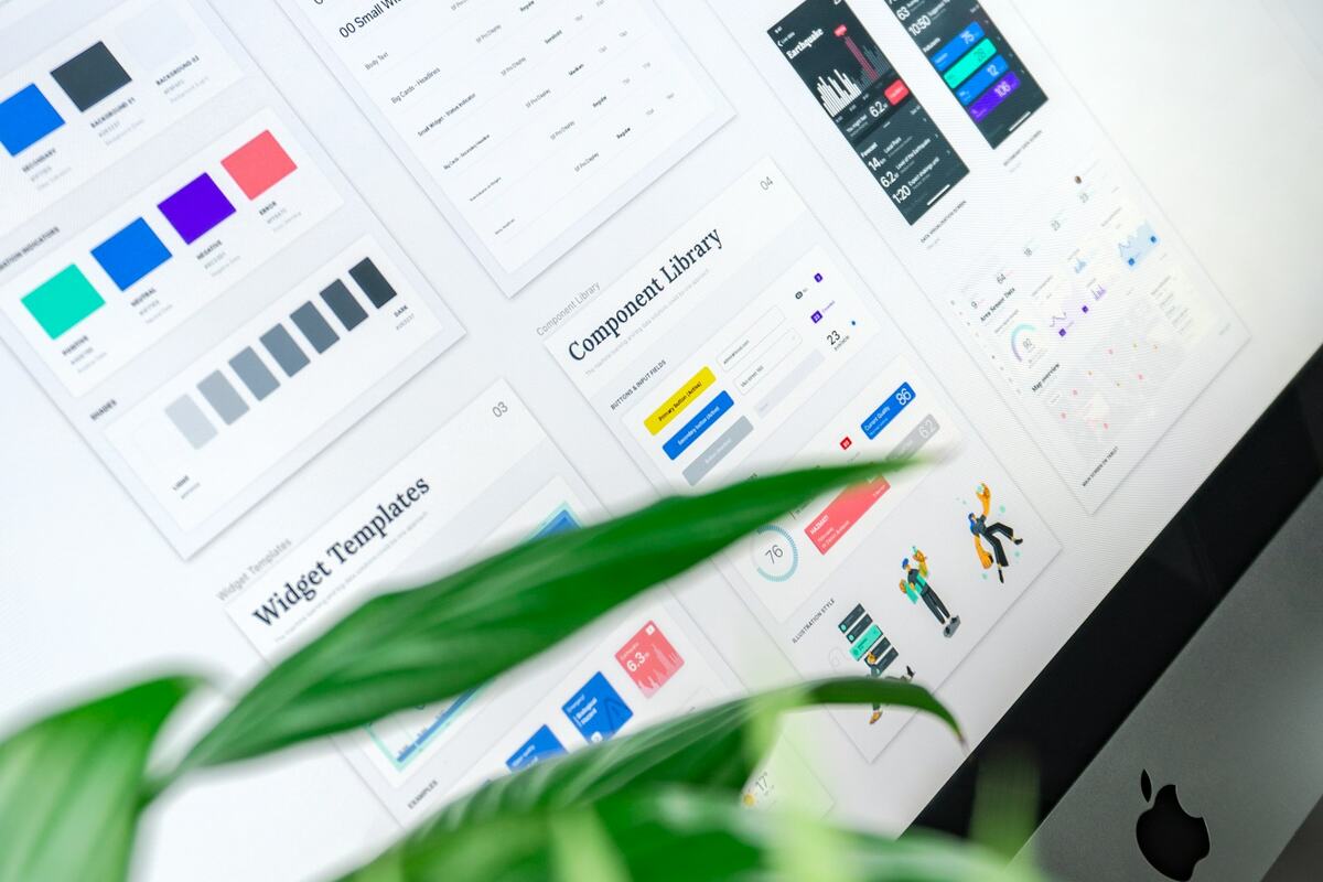 A design system style guide with color swatches and UI components shown on a monitor, with a plant leaf in the foreground